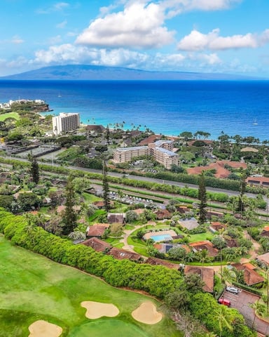 Aerial view of coastal resort community with golf course, red-roofed homes, turquoise ocean, and distant volcanic island