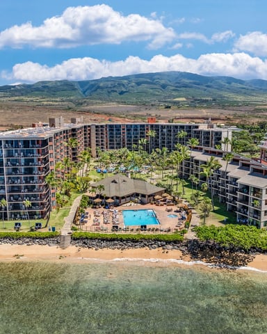 Beachfront resort with multiple apartment buildings, central pool, and sandy beach, with green mountain in background