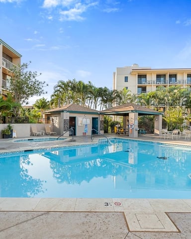 Resort swimming pool with turquoise water, surrounded by multi-story hotel buildings, palm trees, and pool house with teal awning