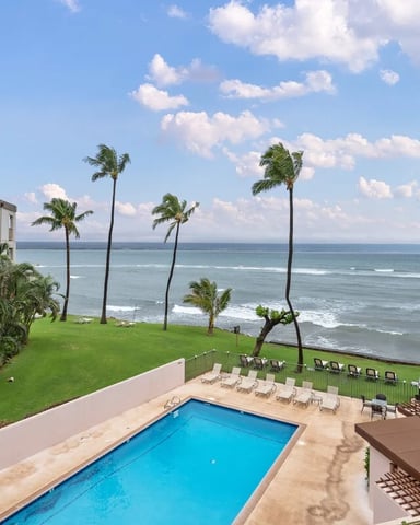 Beachfront resort with swimming pool, palm trees, manicured lawn, and ocean view under blue sky