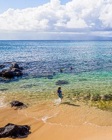 Person wading in clear turquoise water at sandy beach with rocky outcrops and white clouds overhead