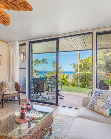 Beachfront living room with ocean view through glass doors, cream sofa, wooden entertainment center, and tropical patio beyond