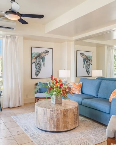Bright tropical living room with blue sofa, round wood coffee table, sea turtle artwork, and ocean view from balcony with palm trees.