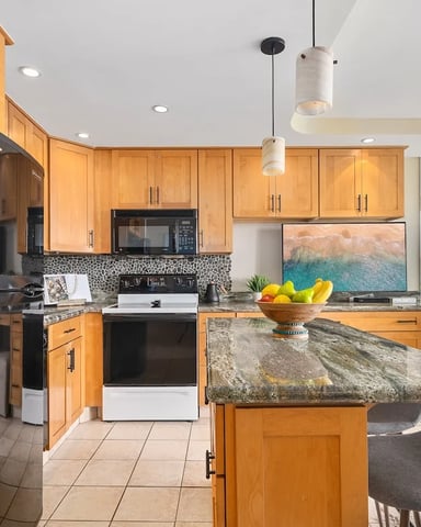 Modern kitchen with honey-wood cabinets, granite island, black appliances, and ocean view through large windows