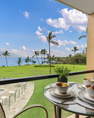 Oceanfront balcony with dining table set for two, overlooking manicured lawn, palm trees, and blue ocean water