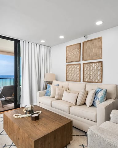 Beachfront living room with cream sofa, armchair, wooden coffee table, and ocean view through sliding glass doors to balcony