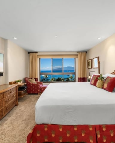 Beachfront hotel bedroom with king bed, ocean view windows, flat-screen TV, and red accent chairs