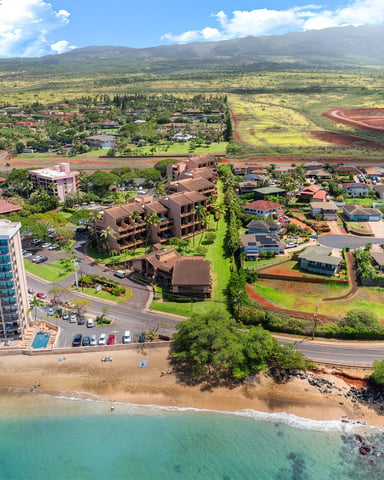 Aerial view of beachfront resort with high-rise hotel, residential neighborhood, and West Maui mountains in background