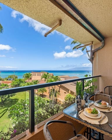 Beachfront lanai with dining table, wicker chairs, and ocean view beyond palm trees and manicured lawn