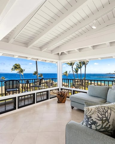 Covered oceanfront lanai with white beamed ceiling, gray seating, and panoramic views of blue ocean, palm trees, and distant coastline.