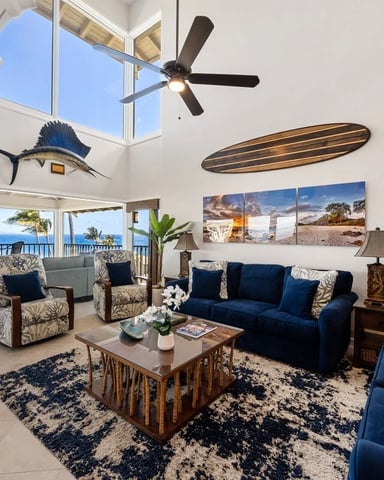 Bright open-plan living room with vaulted ceiling, ocean view, blue sofa, patterned armchairs, and surfboard wall decor
