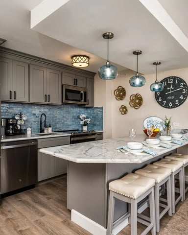 Modern kitchen with gray cabinetry, stainless steel appliances, blue tile backsplash, marble island with seating, and pendant lighting.
