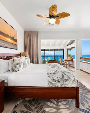Beachfront master bedroom with wooden furniture, ocean view through glass doors, and palm-leaf ceiling fan