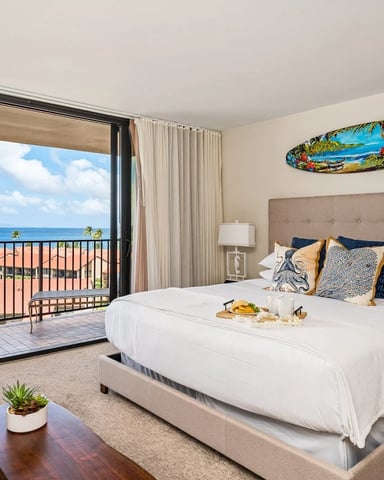 Beachfront bedroom with ocean view from balcony, king bed with taupe headboard, and surfboard wall art