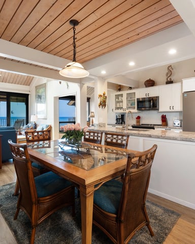 Open-plan kitchen and dining area with ocean view, wood ceiling, white cabinetry, and wooden dining table with ocean-view balcony beyond.