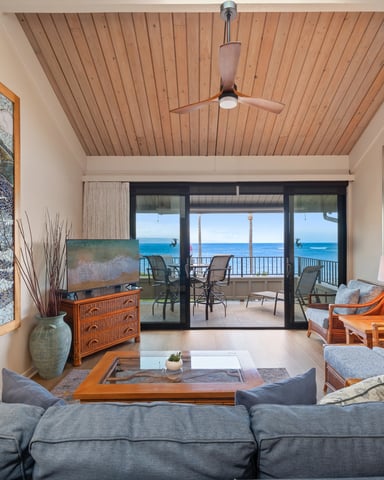 Beachfront living room with gray sofa, wooden furniture, ocean view through glass doors, and ceiling fan