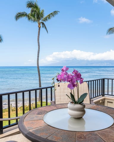 Oceanfront lanai with round table, pink orchid flowers, palm trees, and blue ocean view under clear sky