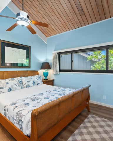 Bedroom with blue walls, wooden ceiling fan, rattan bed, turquoise lamp, and large windows overlooking greenery