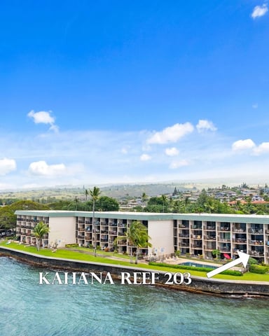 Beachfront condominium building on rocky coastline with turquoise water, palm trees, and distant town and mountains under blue sky