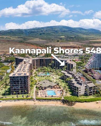 Aerial view of beachfront resort complex with multiple high-rise buildings, pools, and sandy beach, with West Maui mountain in background