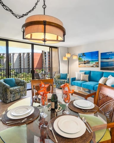 Open-plan living room and dining area with blue sofa, tropical artwork, wood pendant light, and balcony overlooking resort buildings.