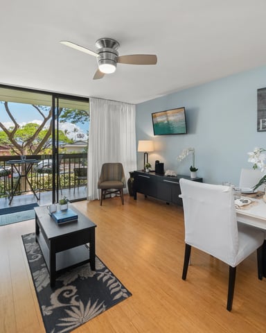 Light-filled living room with blue accent walls, teal sofa, white dining chairs, and ocean view from lanai with large tree visible outside.