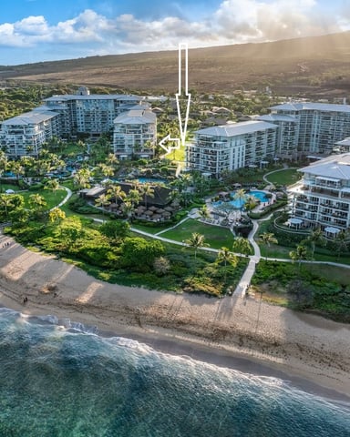 Aerial view of beachfront resort with multiple white buildings, pools, palm trees, sandy beach, and volcanic hillside beyond