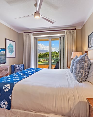 Bedroom with two beds, ocean and mountain view through glass doors, TV on wooden dresser, ceiling fan, and framed artwork.