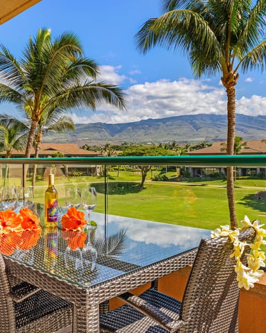 Lanai with woven chairs, dining table with flowers and wine, overlooking manicured lawn, palm trees, and distant mountain under blue sky