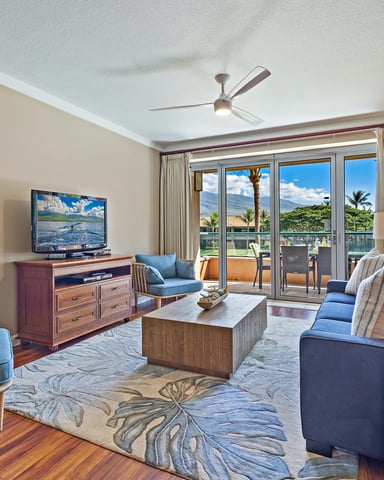 Spacious living room with blue upholstered seating, wooden furniture, ocean and mountain views through glass doors, ceiling fan, and tropical landscape artwork.