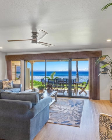 Open-plan living room with gray sectional sofa, ocean view through sliding glass doors, and beachfront patio with dining table.