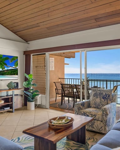 Beachfront condo living room with wood ceiling, ocean view through glass doors, and furnished balcony overlooking water