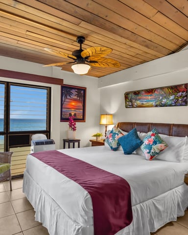 Beachfront bedroom with king bed, tropical decor, wooden ceiling fan, ocean view windows, and wicker seating area