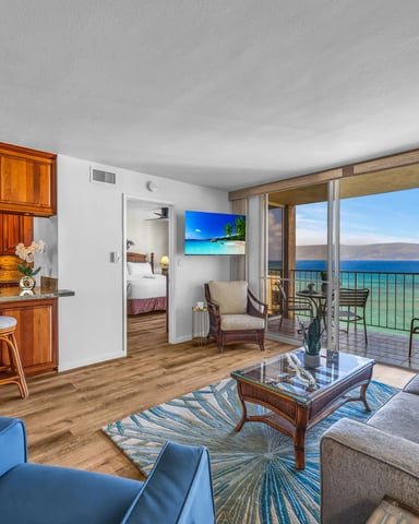 Beachfront condo living room with ocean view, wood cabinetry kitchen, gray sofa, and glass balcony doors overlooking water.