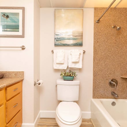 Bathroom with yellow vanity, cork countertop, white toilet, bathtub with cork surround, and coastal artwork on pink walls