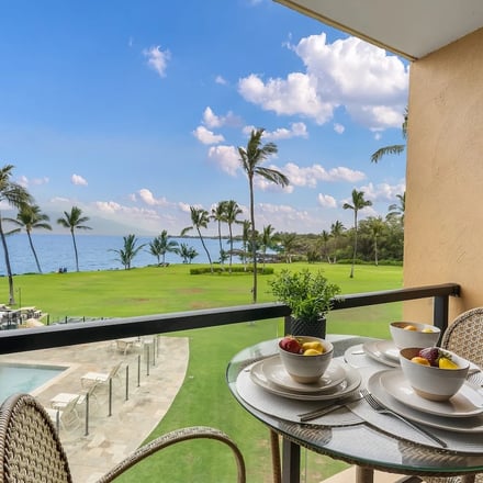 Oceanfront balcony with dining table set for two, overlooking manicured lawn, palm trees, and blue ocean water