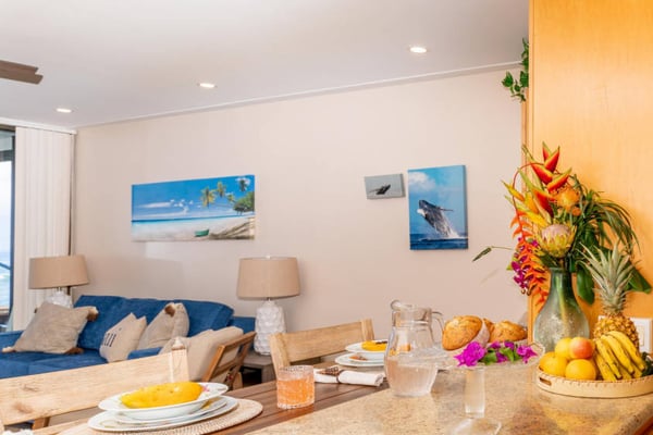 Open-plan living and dining area with blue sofa, dining table set with fruit and place settings, tropical flower arrangement, and beach artwork on walls.