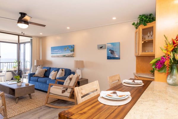 Open-plan living and dining area with blue sofa, wooden dining table, ocean artwork, and balcony access