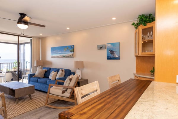 Bright living room with blue sofa, wooden furniture, ocean-view balcony, and coastal artwork on walls