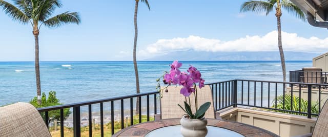 Oceanfront deck with round table, pink orchid plant, palm trees, and clear blue ocean view under sunny sky