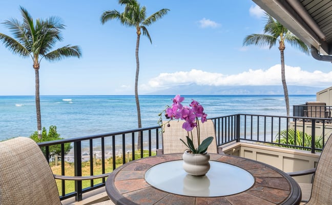 Oceanfront lanai with round table, pink orchids, palm trees, and blue ocean view under clear sky